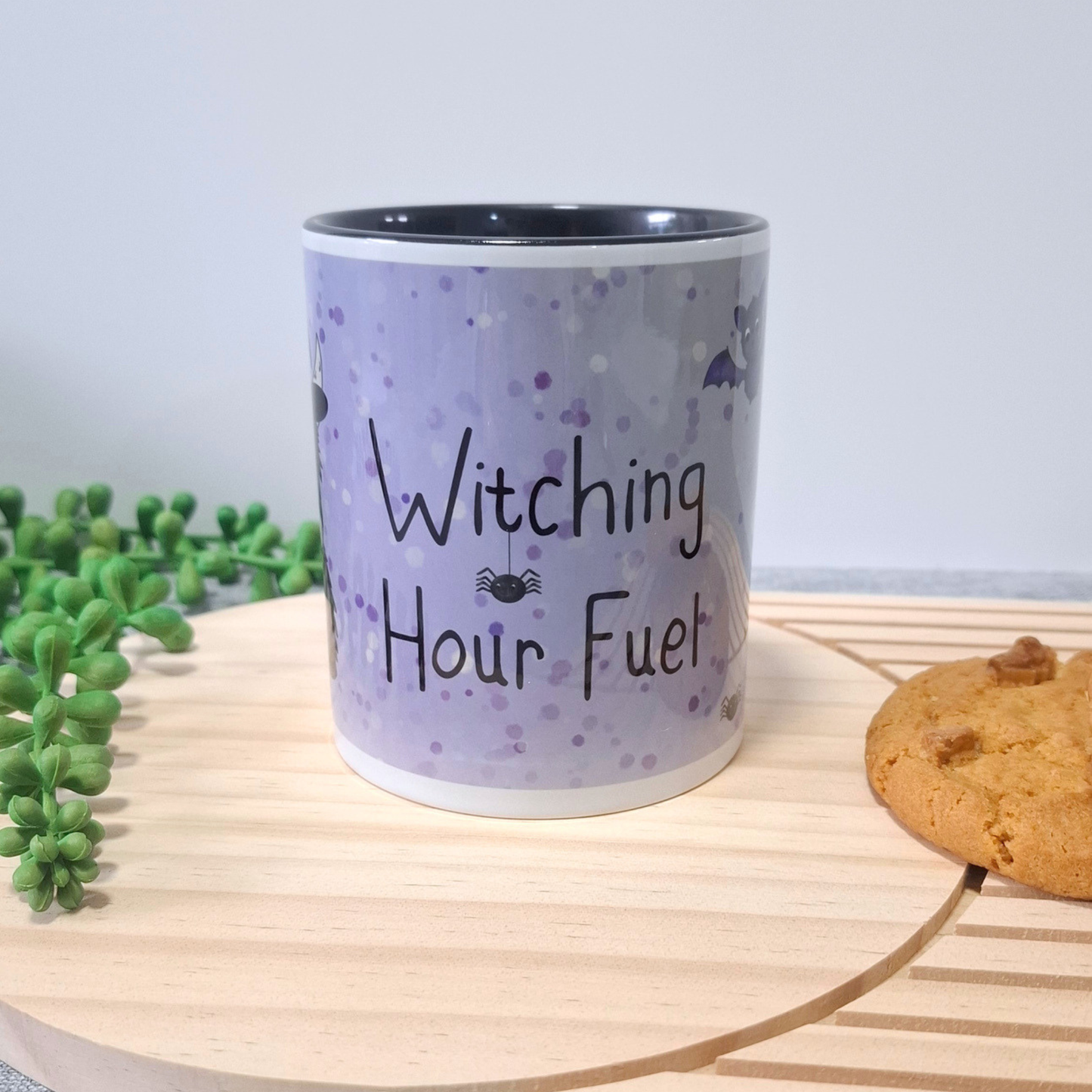 A ceramic mug with a light purple  pattern and the text 'Witching Hour Fuel' in black, styled on a wooden board with a cookie and green plants in the background.