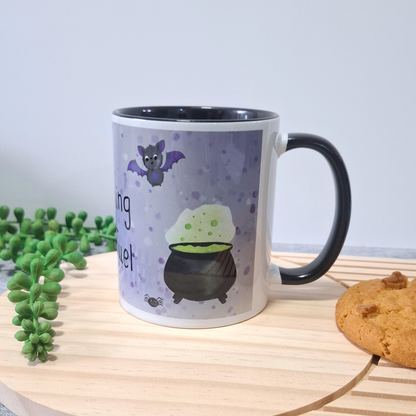 A white ceramic mug with a black handle and interior, decorated with a purple cauldron, green potion, and a bat illustration. The mug is placed on a light wooden board with a green plant and a cookie nearby against a plain light purple background.