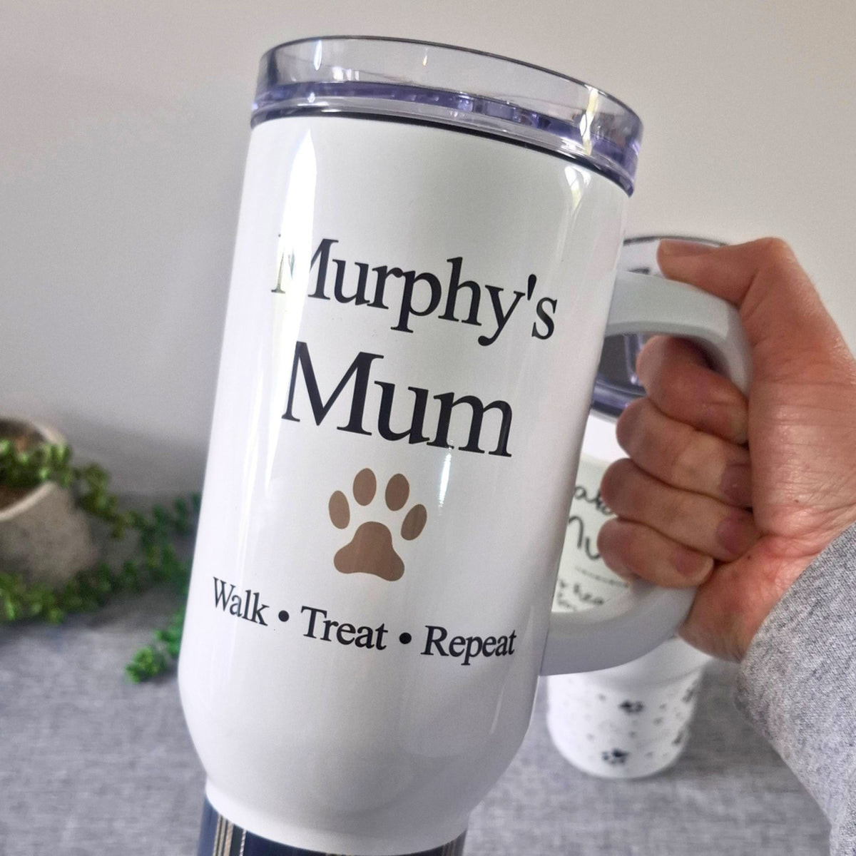 A white insulated travel mug with a clear lid is held by a hand. The mug is printed with the text 'Murphy's Mum' and 'Walk Treat Repeat' with a brown paw print in the center.